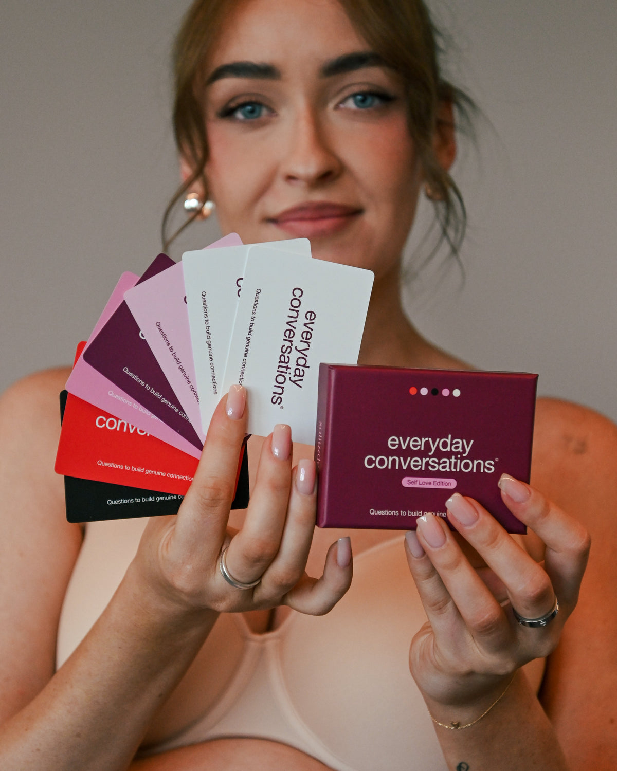 Woman fanning out Everyday Conversations Self Love Edition cards in shades of pink, red and black, with the maroon box in hand